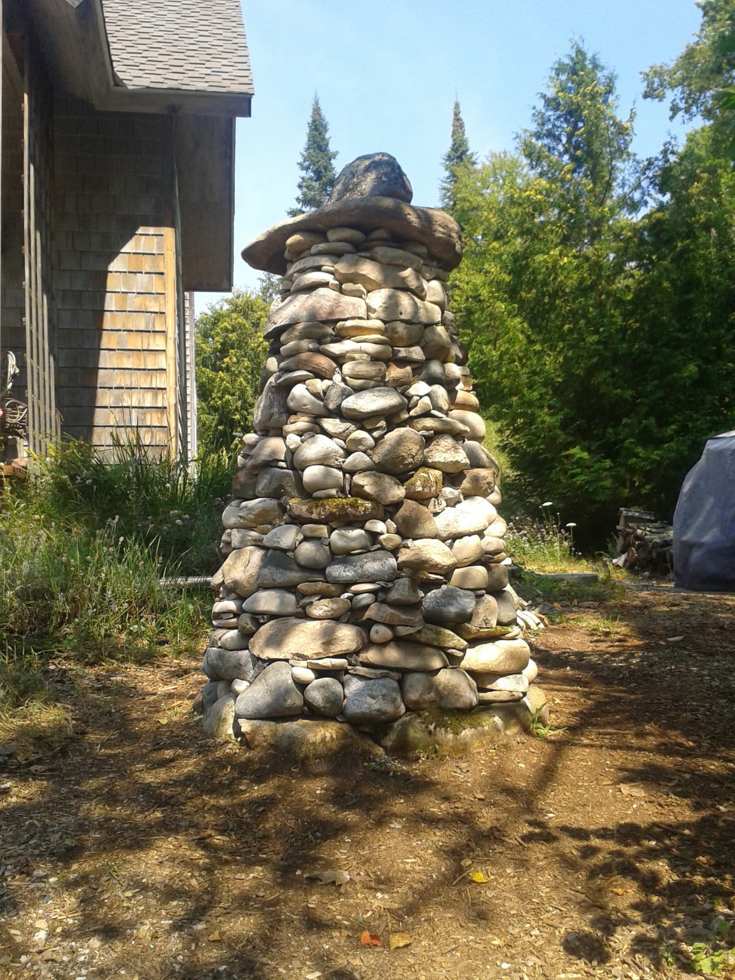 Tall fieldstone cairn with an overhanging capstone beside a shingled house.